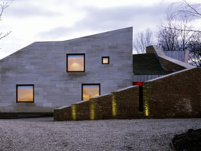 an angular building with a plain white facade on the left with four windows, and a sloped cobblestone area lit by four lights.