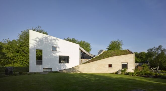 an angular building with a plain white facade on the left with three windows, and a sloped green roof visible on the right. sunlight shines on the building.