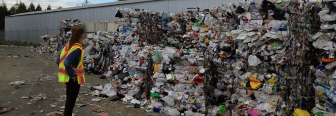 person stands next to piles of plastic at a recycling plant