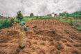 a person dressed in yellow with a tool slung over their shoulder looks out over a field of dirt for planting