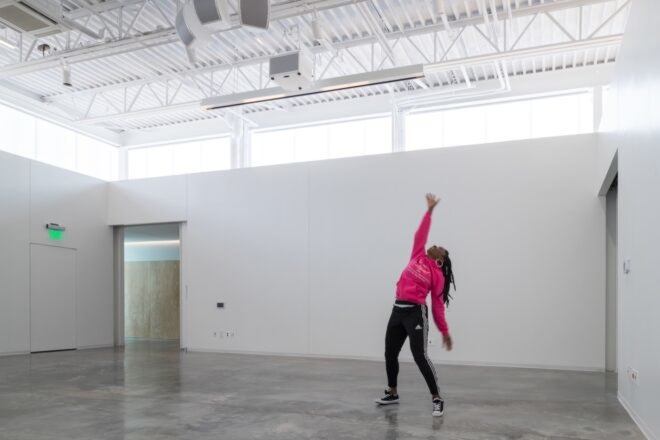 interior studio space with gray floors and white walls. windows line the upper part of the wall, letting in natural light, and a person in a pink top poses with their arm raised toward the ceiling