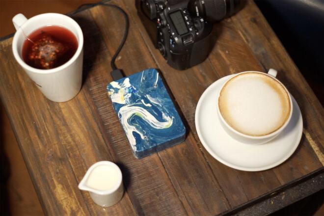a blue and white marbled external charger connected to a camera next to cups of coffee and tea on a wood table