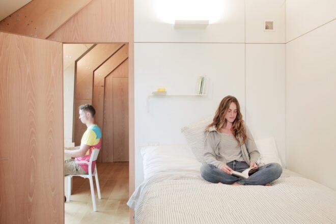 one person in a white bed next to another person at a desk against a glass wall