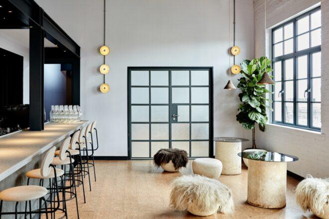 to the left, stools at a bar. to the right, a neutral colored room with pendant lights and tables with backless chairs