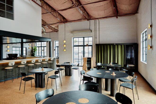the interior of a restaurant with dark colored round tables and chairs, and a bar area with stools