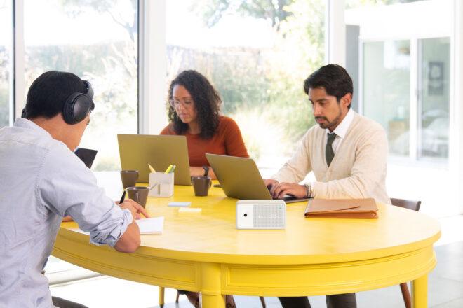 small white air quality monitor on a yellow table, at which three people are working on laptops