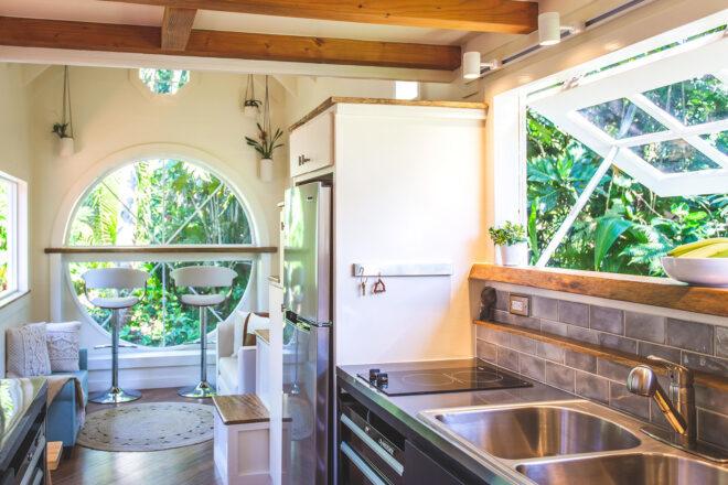 a kitchen area with steel counters and two windows, one rectangular and above the sink, the other circular and on the opposing wall