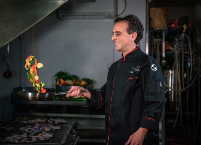 man in black chef's coat flipping vegetables in a pan