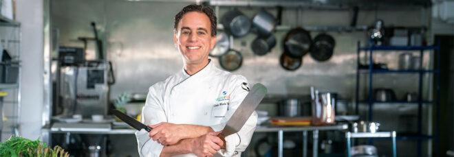 man in white chef coat holding a knife and a spatula near cutting board and cookbooks