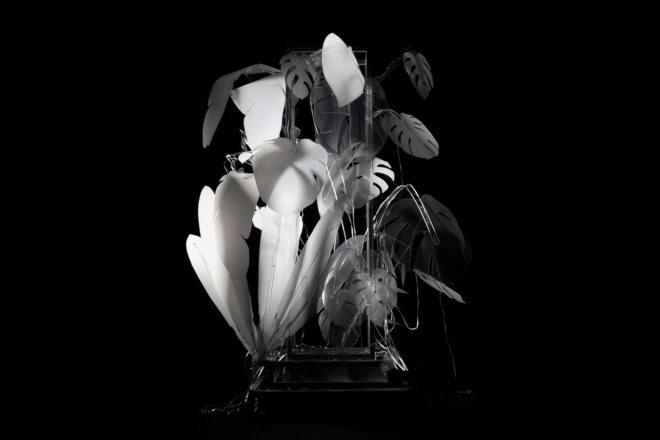 a lit-up white art piece in the shape of a leafy plant against a black background. some leaves in the piece are not lit-up, which signifies rainforest loss