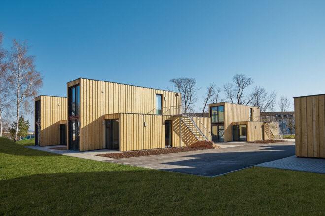 a grassy area surrounds several shipping container apartments covered in timber