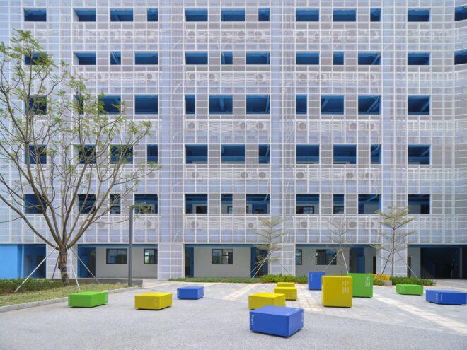 a school building with a courtyard in front of it. blue, yellow and green seating areas are placed throughout the courtyard