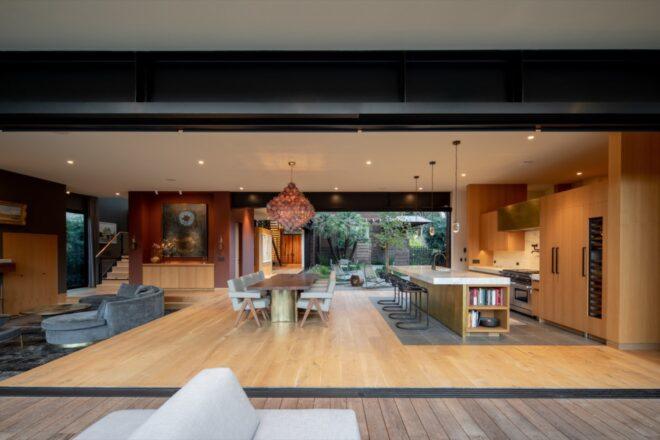 the interior of a modern home with wood accents and artistic ceiling lights overhanging the dining room and kitchen. the opposite wall has large open doors to expose the woods outside