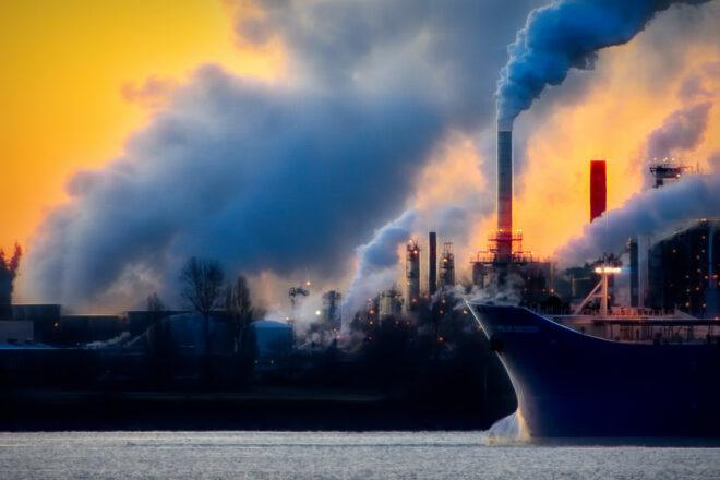a black ship on water with a factory behind it emitting gas into the air