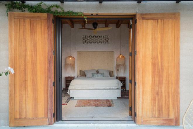an open wood doorway that shows the interior of a bedroom with white bedding and hanging lights over the bedside tables