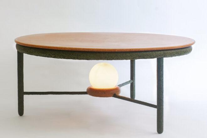 a coffee table with a brown top and dark green accents and legs. underneath the table is a lit round lightbulb.