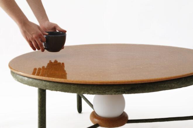 a coffee table with a brown top and dark green accents and legs. underneath the table is an attached, round lightbulb, unlit. a pair of hands are setting down a dark coffee mug on the table.