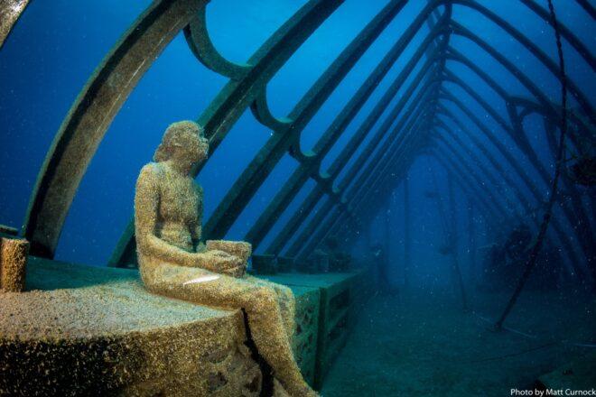 statue of person sitting on a bench in an underwater greenhouse