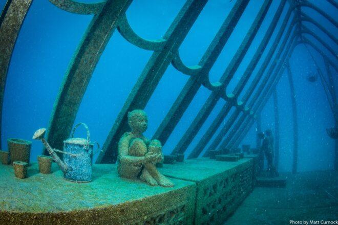 statue of child sitting near a watering can in an underwater greenhouse