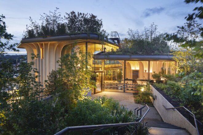 wood-framed building with glass walls and lush green roofs lit from within at night