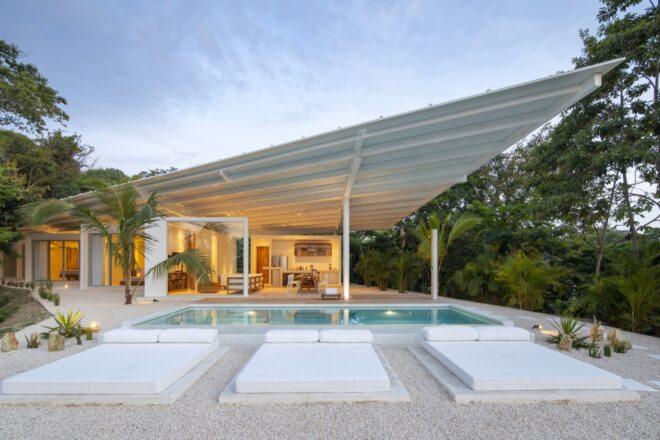 a white home with an angular roof lit up from the inside. in front of the home is a pool with three bed-like seating areas
