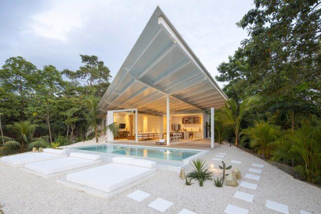 a white home with an angular roof lit up from the inside. in front of the home is a pool with three bed-like seating areas. to the side of the pool is a white stone walkway and plants
