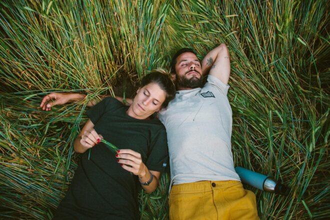 One person in green T-shirt dress and other person in white shirt and yellow shorts both lying down in tall grass