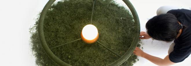 a coffee table made of grass clippings with attached, lit-up, round lightbulb. a person to the right of the table touches the table's outer edge.