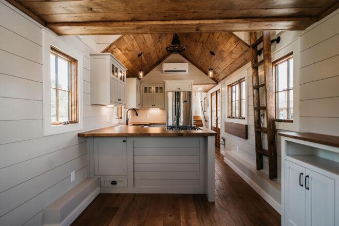 interior of a tiny home with white cabinets and wood ceilings and accents in the kitchen. a ladder is mounted on the wall