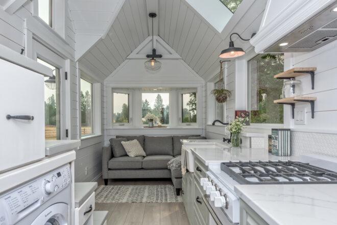 the interior of a tiny home featuring white shiplap walls, a sofa near a bay window and a kitchen