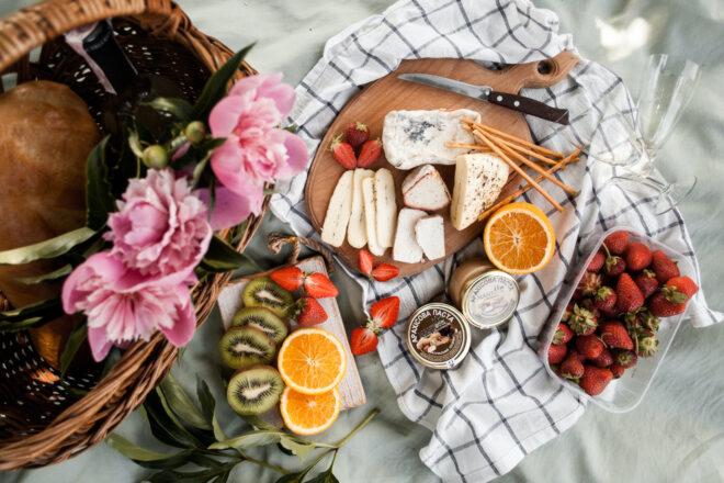 wood board with cheeses and fruit near wicker basket on plaid blanket