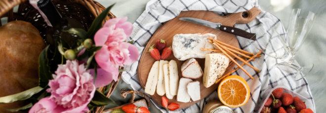 wood board with cheeses and fruit near wicker basket on plaid blanket