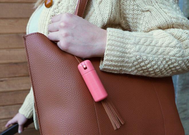 an arm in a white sweater clutching a brown purse with a pink bottle of sanitizer attached