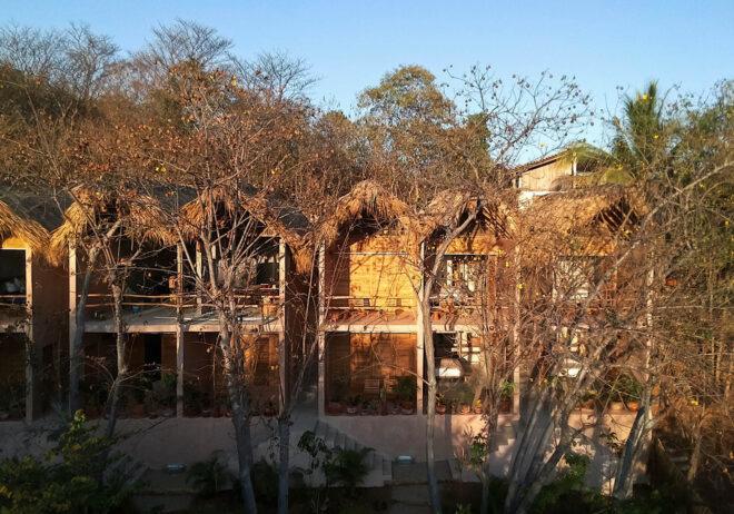 a row of six hotel buildings with fringed, pitched rooftops, partially obscured by bare trees in front of the buildings.