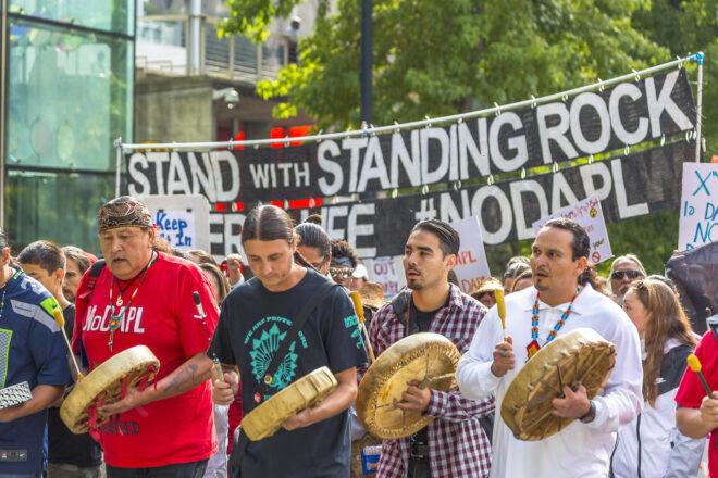 a group of four people stand in front of a crowd and hold drums, with a sign in the background reading "Stand with Standing Rock"