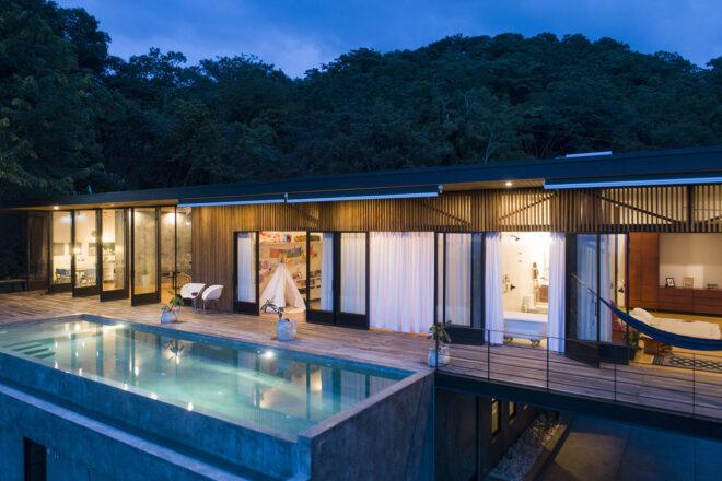 a long rectangular home shot from an angle, with a back porch with a wood deck with a pool. it is dark outside and the home is lit from within.
