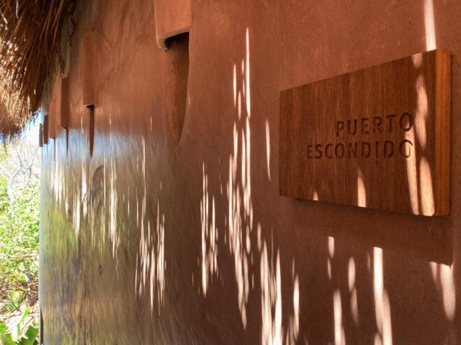 a dusty pink wall with dappled sunlight and a sign reading "Puerto Escondido."