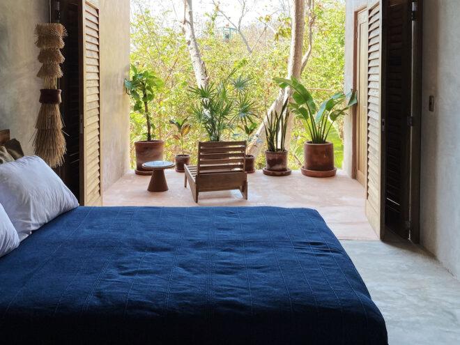 a bedroom with blue sheets and white pillows. in front of the bed, a set of wood doors open to a terrace with a chair and potted plants that overlook a green jungle.
