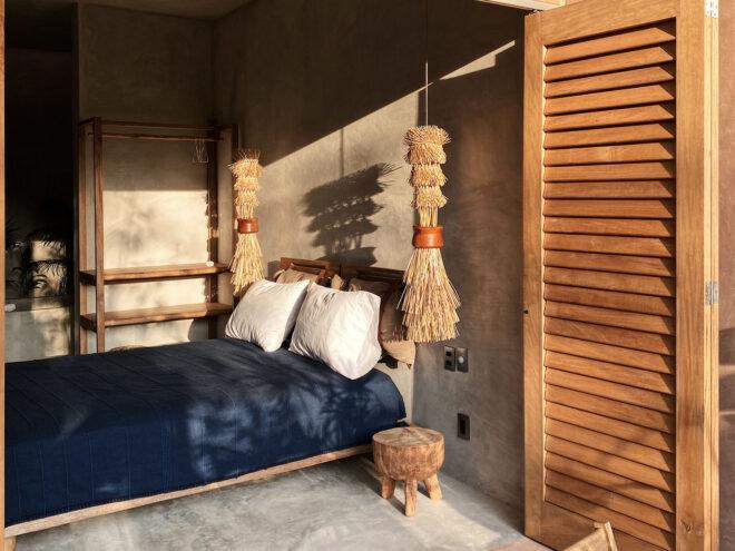 a bedroom with natural light filtering in onto a bed with blue sheets and white pillows. a round bedside table sits underneath a hanging, fringed decoration that matches with a similar object on the other side of the bed.