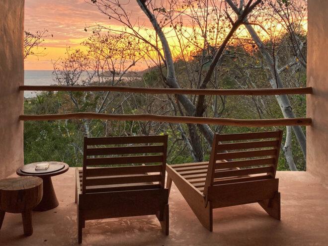 two wood chairs and two short round tables to the left in front of two bars that protect a terrace overlooking the sunset in the jungle.
