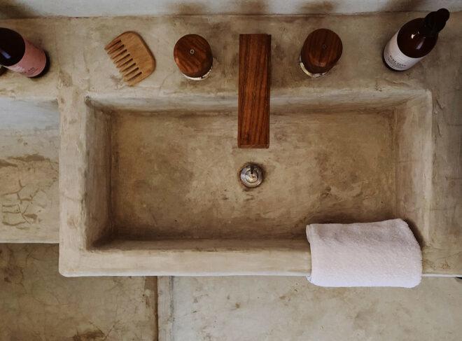 a grey cement-like square sink with wood fixtures. two bottles, a comb and a white hand towel sit around the sink.