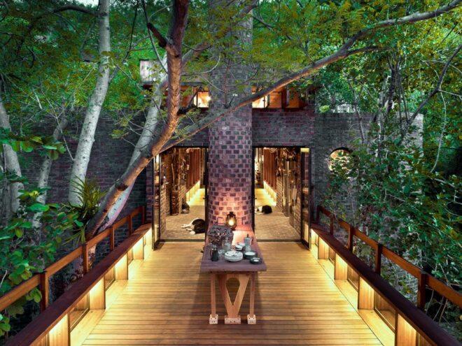 a lit up wood bridge connecting to a brick house, with a brick pillar in the middle of it. trees surround the structure.