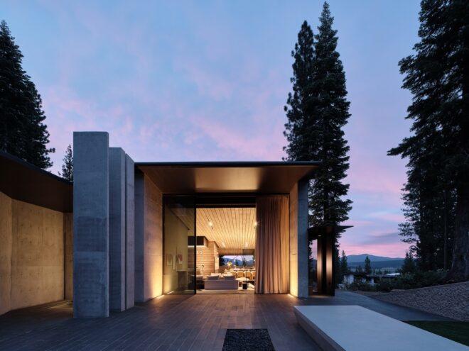 a block-like structure with a full-height sliding glass door entrance showing warm light and wood accents within the home. in the background is a purple and blue horizon.
