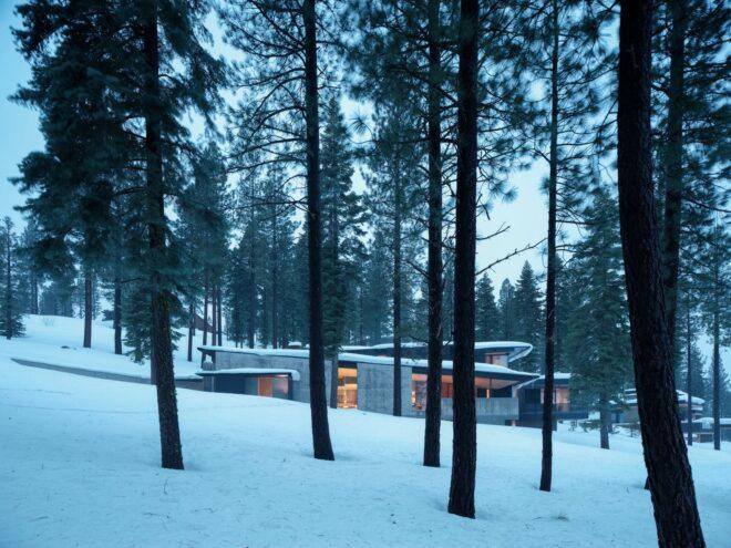 a home with a gray facade dotted with windows. the home is on a snowy slope in the middle of a forest.