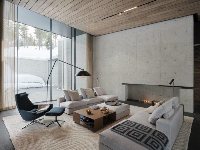 a neutral-toned living room with two off-white couches with pillows, a dark chair with matching foot rest, a white ottoman next to a brown coffee table, black floor lamp and fireplace built into the gray wall. to the left are glass panels that lok out onto a patio area covered in snow.