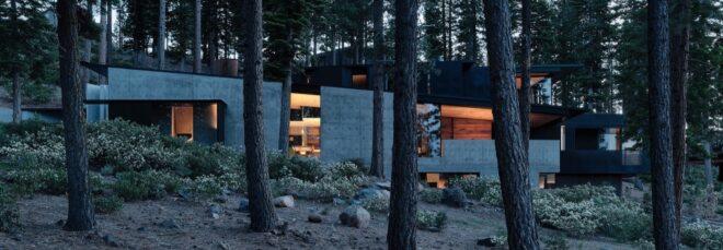 a home with a gray facade nestled within a slope in a forest. warm light shines from within the home.