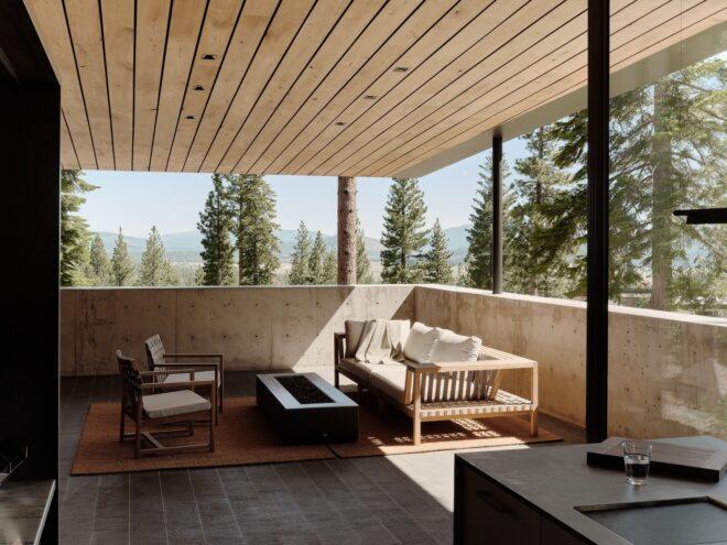 a covered outdoor patio area with a dark coffee table placed between a wood sofa bench with white cushions and two wood-frame chairs. the patio looks out on a forest.