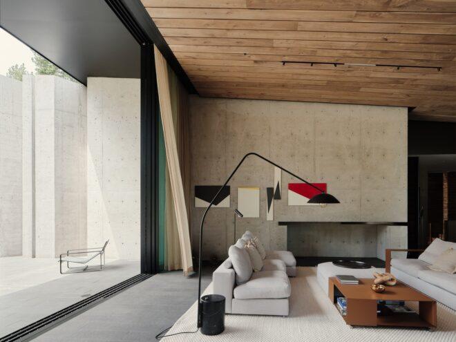 to the left, a floor-to-ceiling window showing an outdoor sitting area with one chair. to the right, a neutral-toned living room with two off-white couches with pillows, a dark chair with matching foot rest, a white ottoman next to a brown coffee table, black floor lamp and fireplace built into the gray wall. five pieces of various-sized black, white and red abstract art hang on the wall.