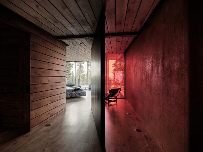 a hallway split in half by a divider. to the left are wood accents leading to a bedroom with floor-to-ceiling windows. to the right is a sitting area lit up red with colored glass.