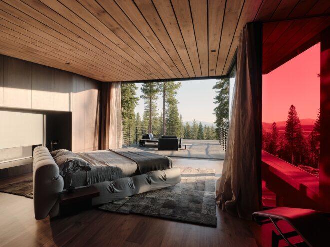 to the left, a bedroom with a neutral-toned bed placed in the middle of the room on a neutral-toned rug. past the bed is an open area leading out to a deck with chairs that overlook the forest outside.. to the right is a sitting area lit up red with colored glass.